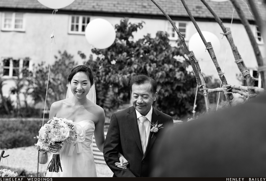 A beaming bride smiles at her groom down the aisle as she walks up the garden path of River Cottage on her fathers arm before her wedding