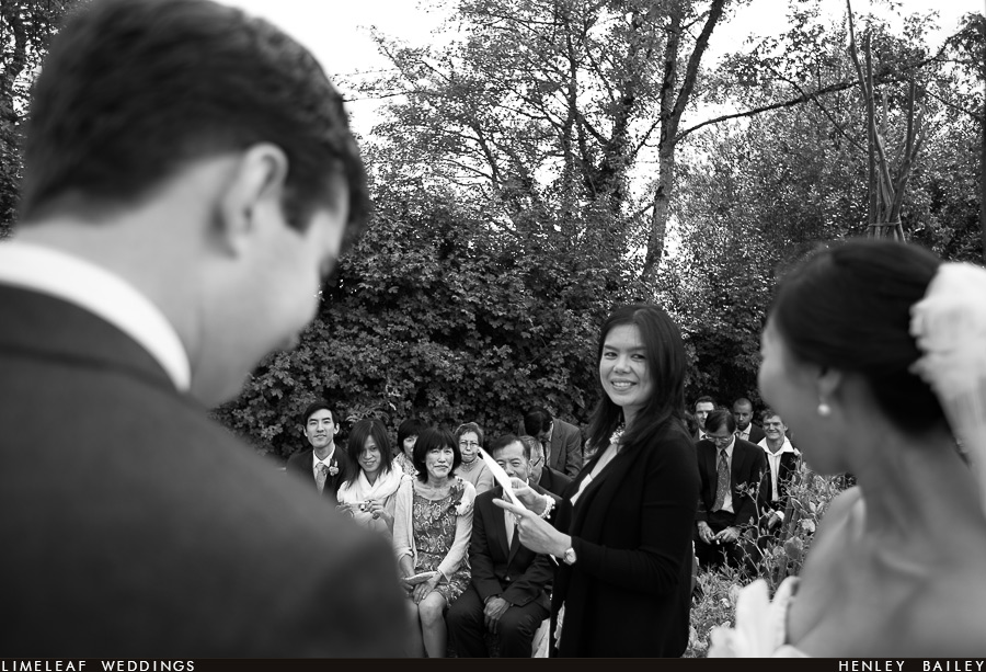 A friend smiles at the bride and groom while their guests look on as she recites a reading at their River Cottage garden wedding